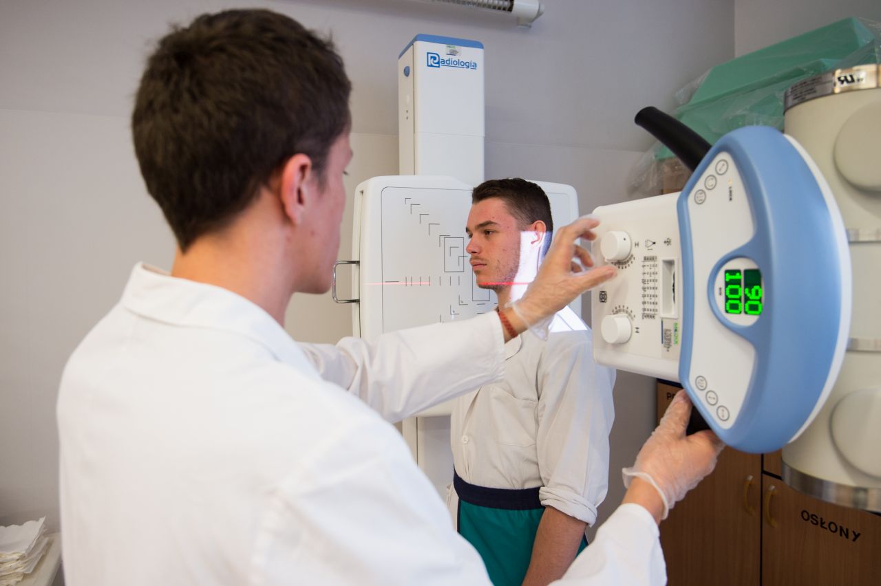 Zdjęcie przedstawia dwóch uczniów uczących się wykonywania badania diagnostycznego. Jeden z uczniów ustawia aparat rentgenowski do wykonania badania, drugi uczeń występuje w roli pacjenta stojącego przy aparacie.
