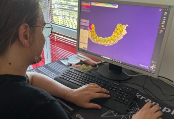 Zdjęcie przedstawia słuchacza szkoły wykonującego komputerową obróbkę pracy protetycznej. Słuchacz siedzi przed komputerem. Na monitorze widać program do prac protetycznych 