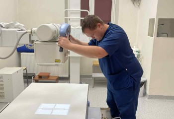 Zdjęcie przedstawia słuchacza wykonującego badanie diagnostyczne z zakresu elektrokardiologii. Widzimy aparat rentgenowski i dotykającego go słuchacza