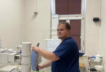 Zdjęcie przedstawia słuchacza wykonującego badanie diagnostyczne z zakresu elektrokardiologii. Widzimy aparat rentgenowski i dotykającego go słuchacza