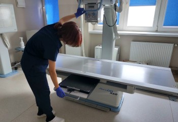 Zdjęcie przedstawia słuchacza wykonującego badanie diagnostyczne z zakresu elektrokardiologii. Widzimy aparat rentgenowski i dotykającego go słuchacza